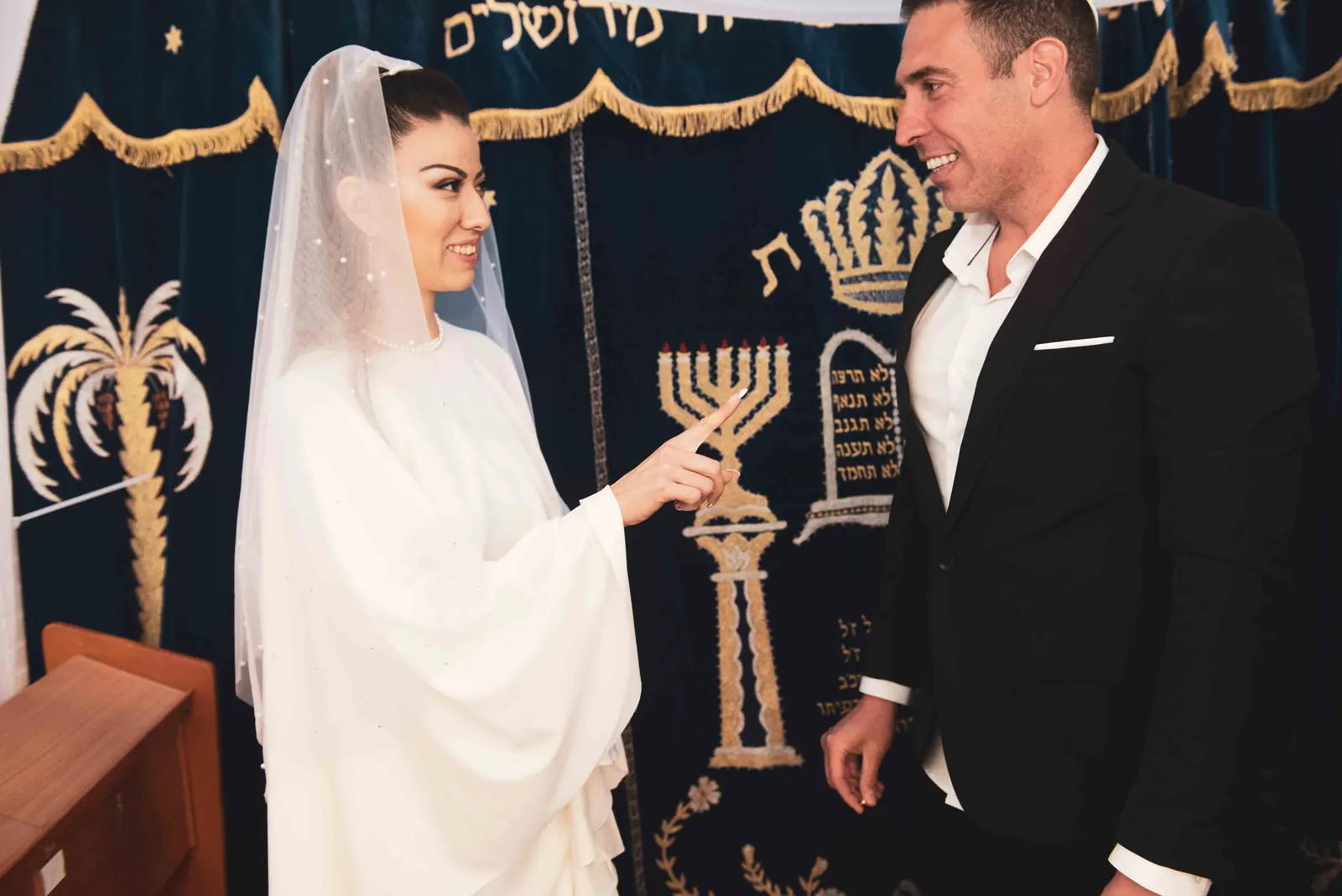 כלה מצביעה על פרוכת בית הכנסת בזמן שהחתן מחייך